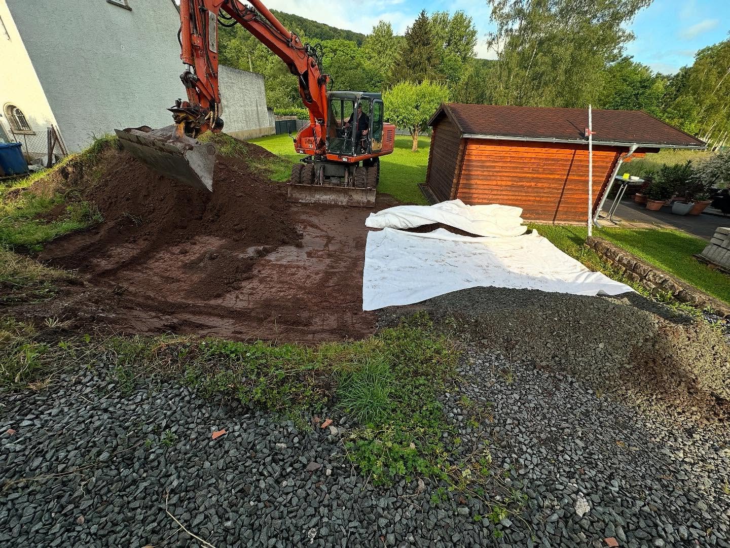 Mobilbagger schiebt Mutterboden zu einem Wall auf, daneben liegt weißes Trennvlies über der frisch geschütteten Schotterfläche vor einem Holz-Gartenhaus.