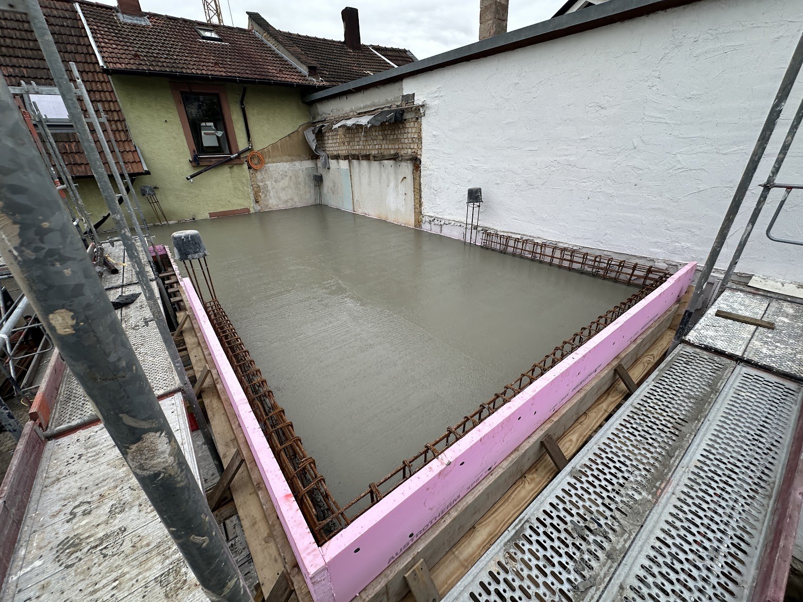 Frisch betonierte und geglaettete Decke mit pinker Austrotherm-Randdaemmung, Anschlussbewehrung und Deckenstuetzen zwischen zwei Altbau-Brandwaenden