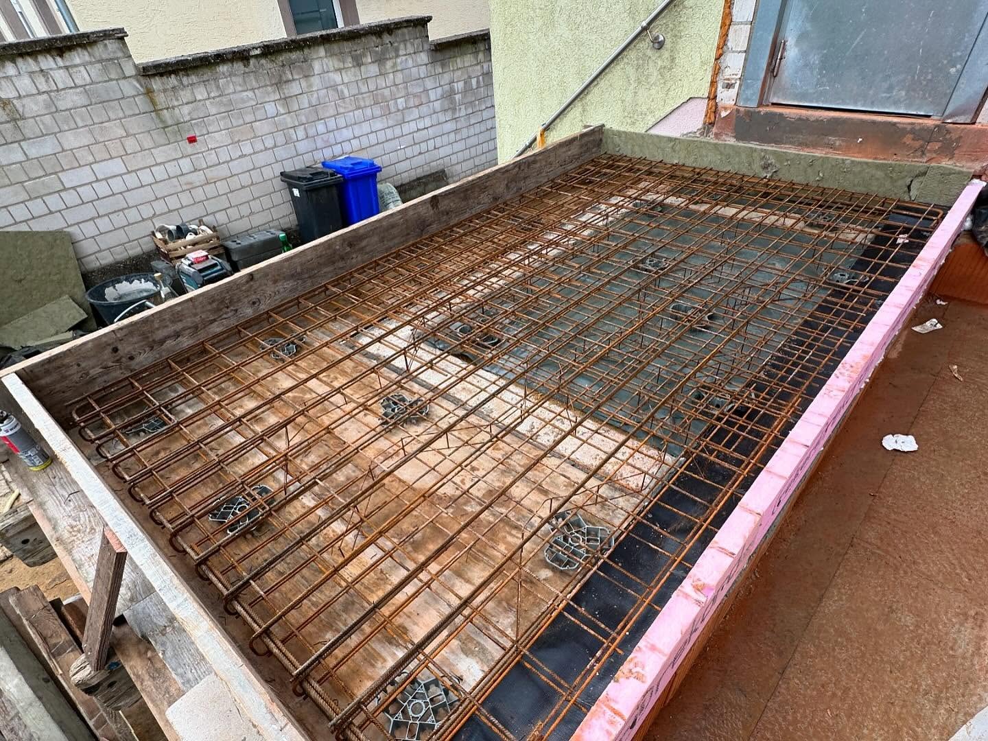 Fertig verlegte, zweilagige Baustahlmatten-Bewehrung mit Abstandhaltern und Randkoerben in der Holzschalung der Bodenplatte auf schwarzer Trennfolie, umlaufend pinke Randdaemmung, kurz vor dem Betonieren.