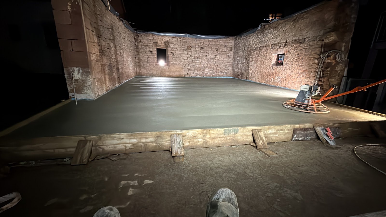 Nachts unter Baustrahler frisch eingebrachte und fluegelgeglaettete Betonbodenplatte innerhalb alter Sandstein-Umfassungsmauern, rechts die Betonglaettmaschine