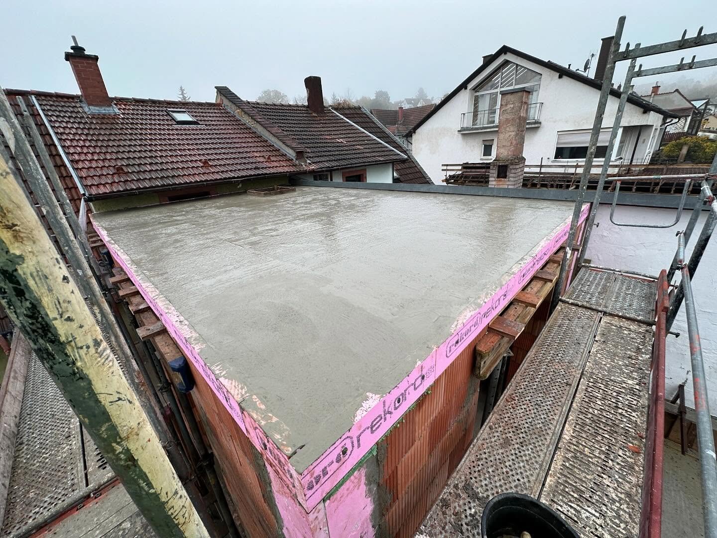 Frisch betonierte, glatt abgezogene Geschossdecke des Anbaus aus der Gerueststellung fotografiert, mit rosa Randdaemmstreifen und Schalungstraegern an der Deckenkante, dahinter Altbaudaecher im Nebel