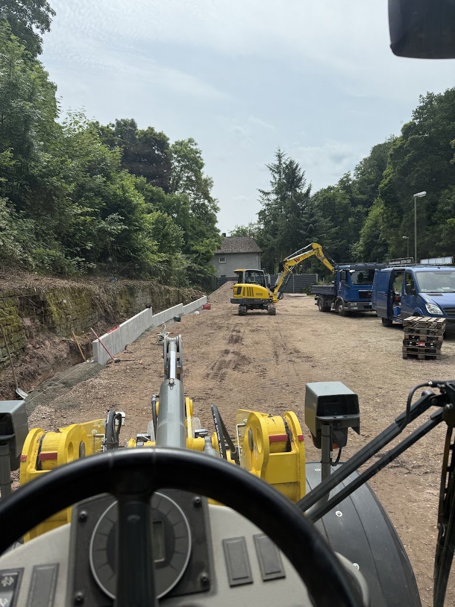 Blick aus der Baggerkabine über Lenkrad und gelbe Hydraulikarme auf eine Baustelle mit frisch gesetzten weißen Betonwinkel-Stützelementen an der Böschung, gelbem Mobilbagger, blauem Kipper und dem eigenen Firmentransporter.