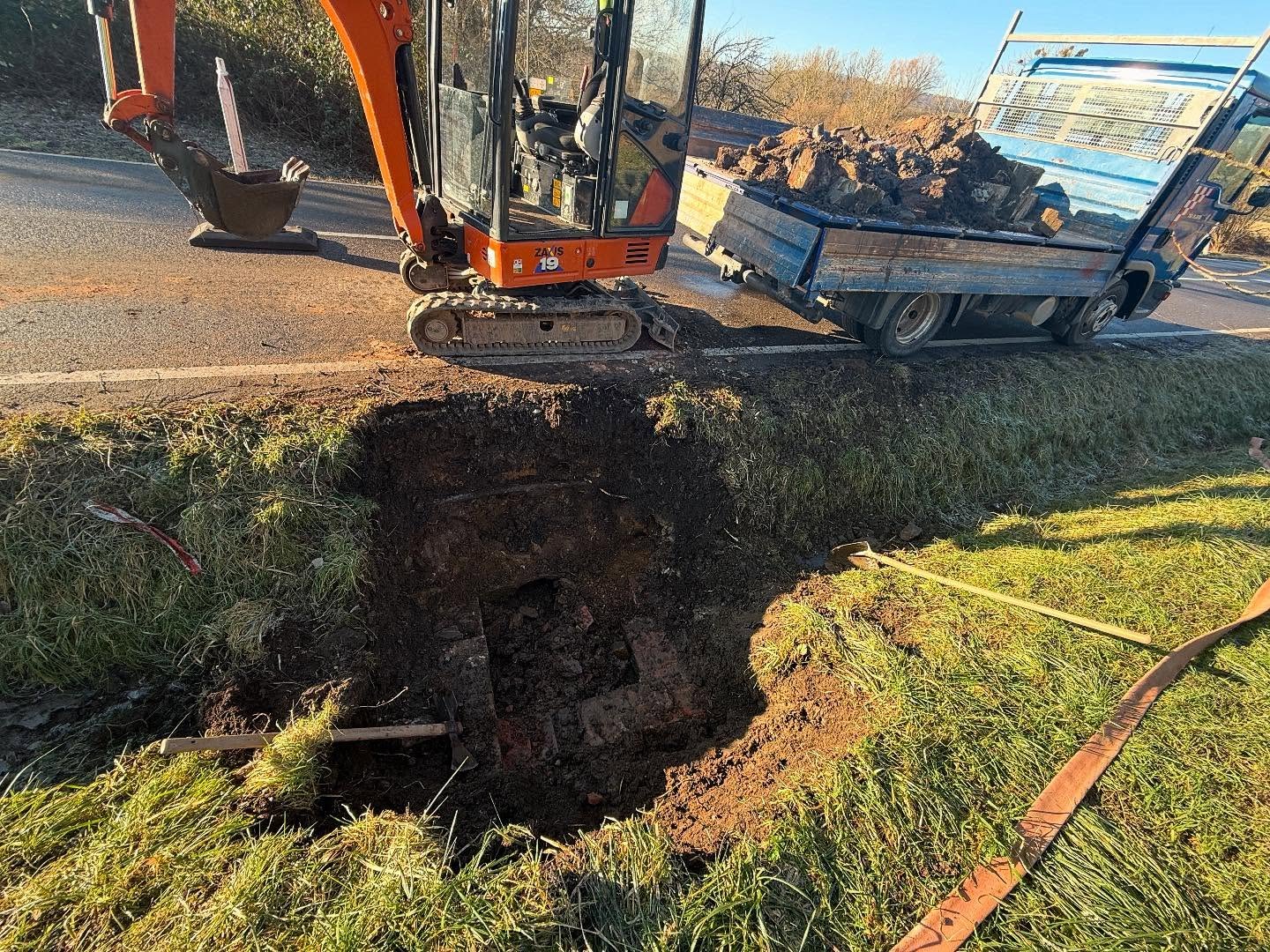 Oranger Hitachi-Minibagger hebt von der Fahrbahn aus die Baugrube für den Einlaufschacht in der Grasböschung aus, daneben der bereits mit Aushub beladene blaue Kipper.