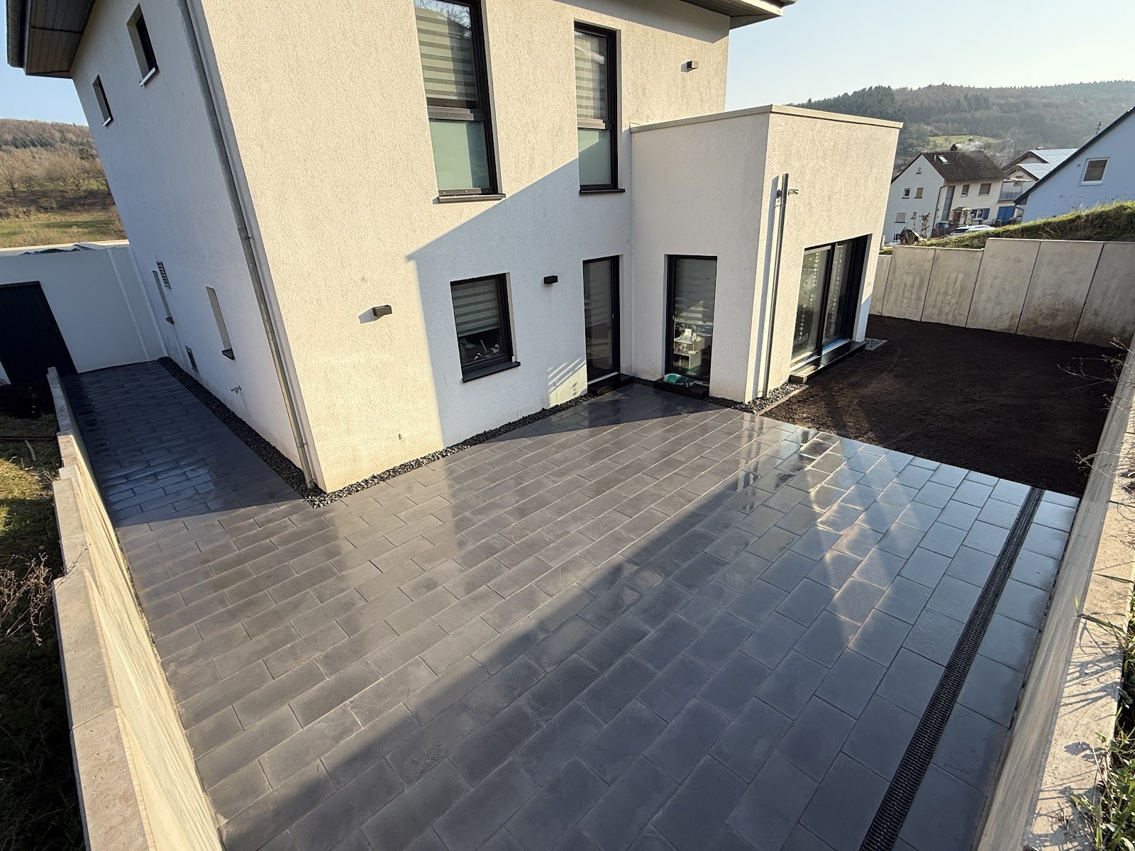 Frisch gepflasterte graue Terrassenflaeche mit Entwaesserungsrinne und Betonmauer-Einfassung an einem hellen Neubau mit anthrazitfarbenen Fenstern