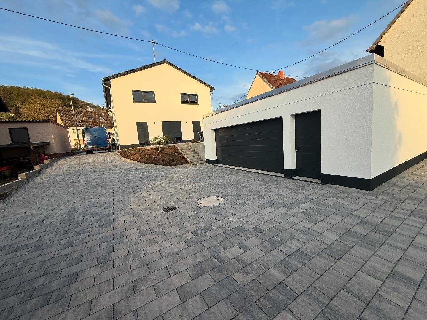 Grossflaechiger grau melierter Pflasterhof mit Entwaesserungsrinne und Schachtdeckel vor einer weissen Doppelgarage mit Flachdach, dahinter der cremefarbene Neubau mit Aussentreppe und L-Stein-Beet