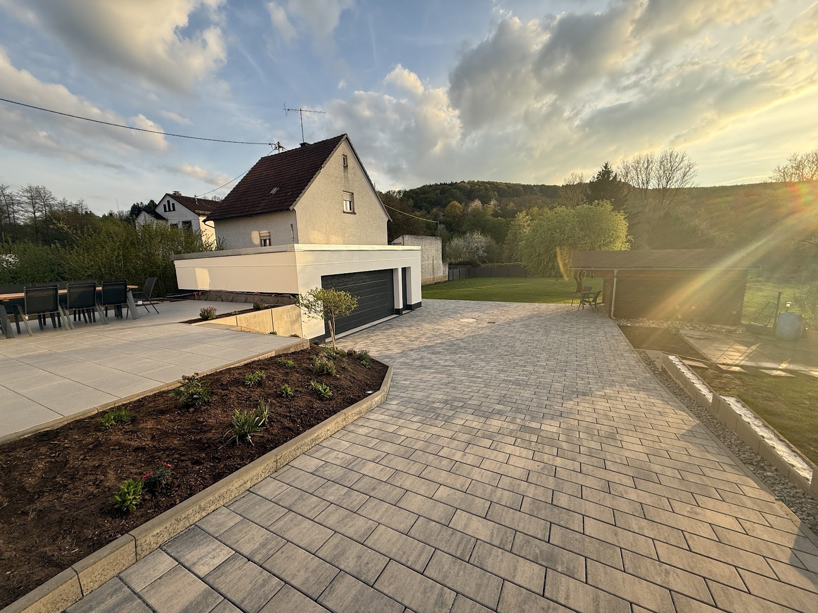 Weitlaeufiger gepflasterter Hof in Grautoenen mit angrenzender Plattenterrasse, bepflanztem Beet und Garagenanbau im Abendlicht vor bewaldeten Huegeln