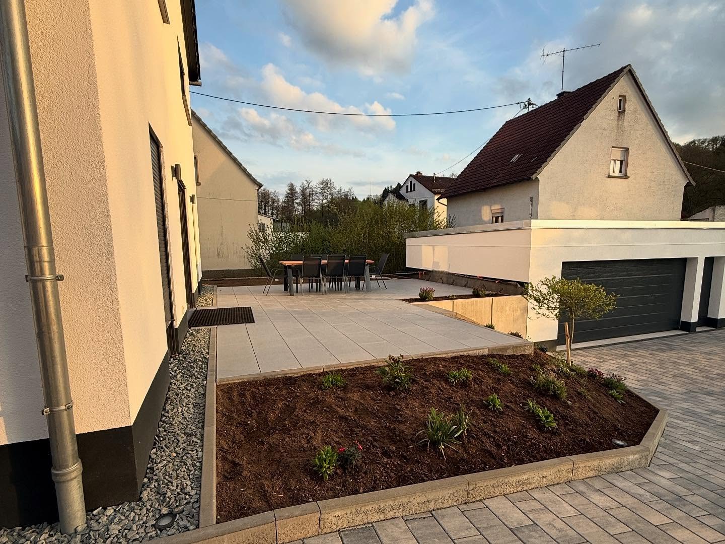 Grossformatige helle Betonplatten-Terrasse mit Esstisch und sechs Stuehlen entlang der Hausseite, davor ein frisch bepflanztes Beet in Randsteineinfassung mit Kugelbaeumchen, rechts die Doppelgarage und die anschliessende Pflasterflaeche
