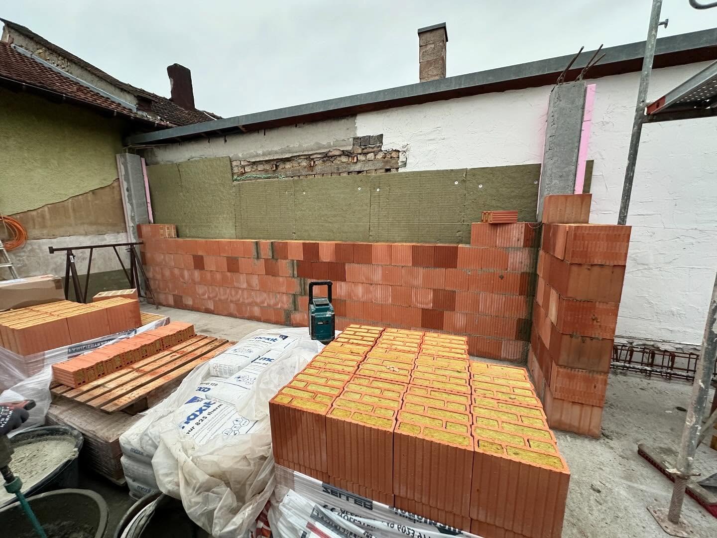 Halbhoch aufgemauerte Poroton-Planziegelwand des Anbaus vor der gedaemmten Bestandsfassade, davor Paletten mit wollgefuellten Waermedaemmziegeln und Maxit-Moertelsaecken.