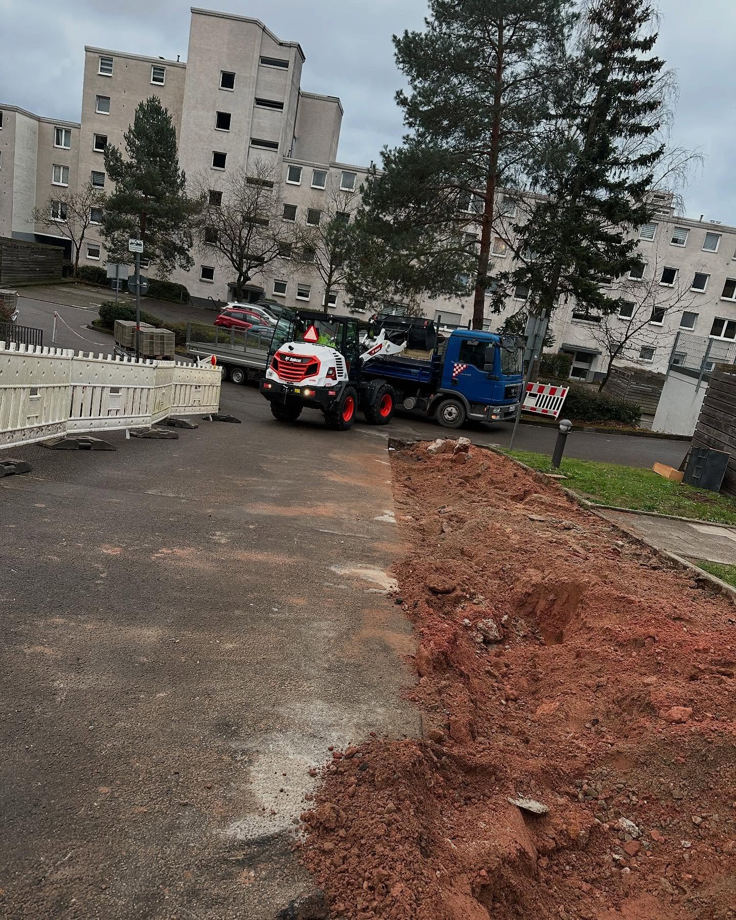 Baustellenübersicht in einer Wohnsiedlung: ein weißer Bobcat-Radlader belädt einen blauen Kipper-LKW, rechts ein offener Erdgraben mit rotem Aushub, links weiße Kunststoff-Absperrgitter.