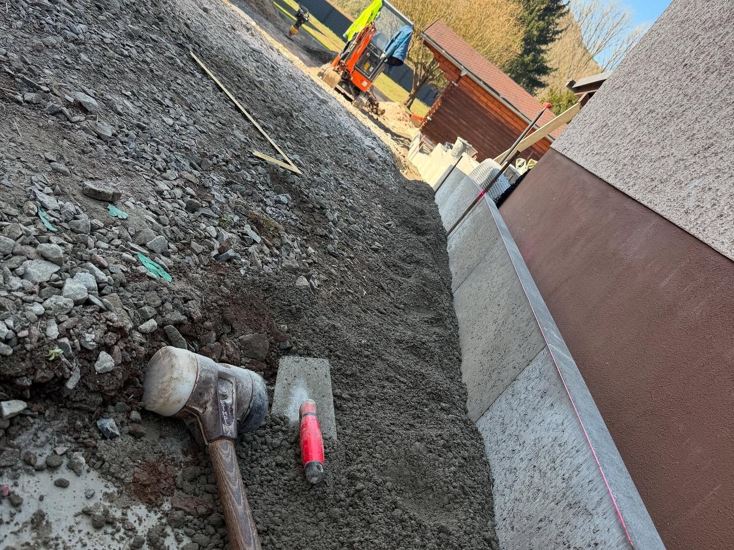Nahaufnahme entlang einer frisch gesetzten Betonrandstein-Kante mit gespannter Richtschnur, im Erdreich liegen ein Gummihammer und eine rote Markierungsspraydose, links aufgeschuetteter Schotter, hinten ein oranger Bagger