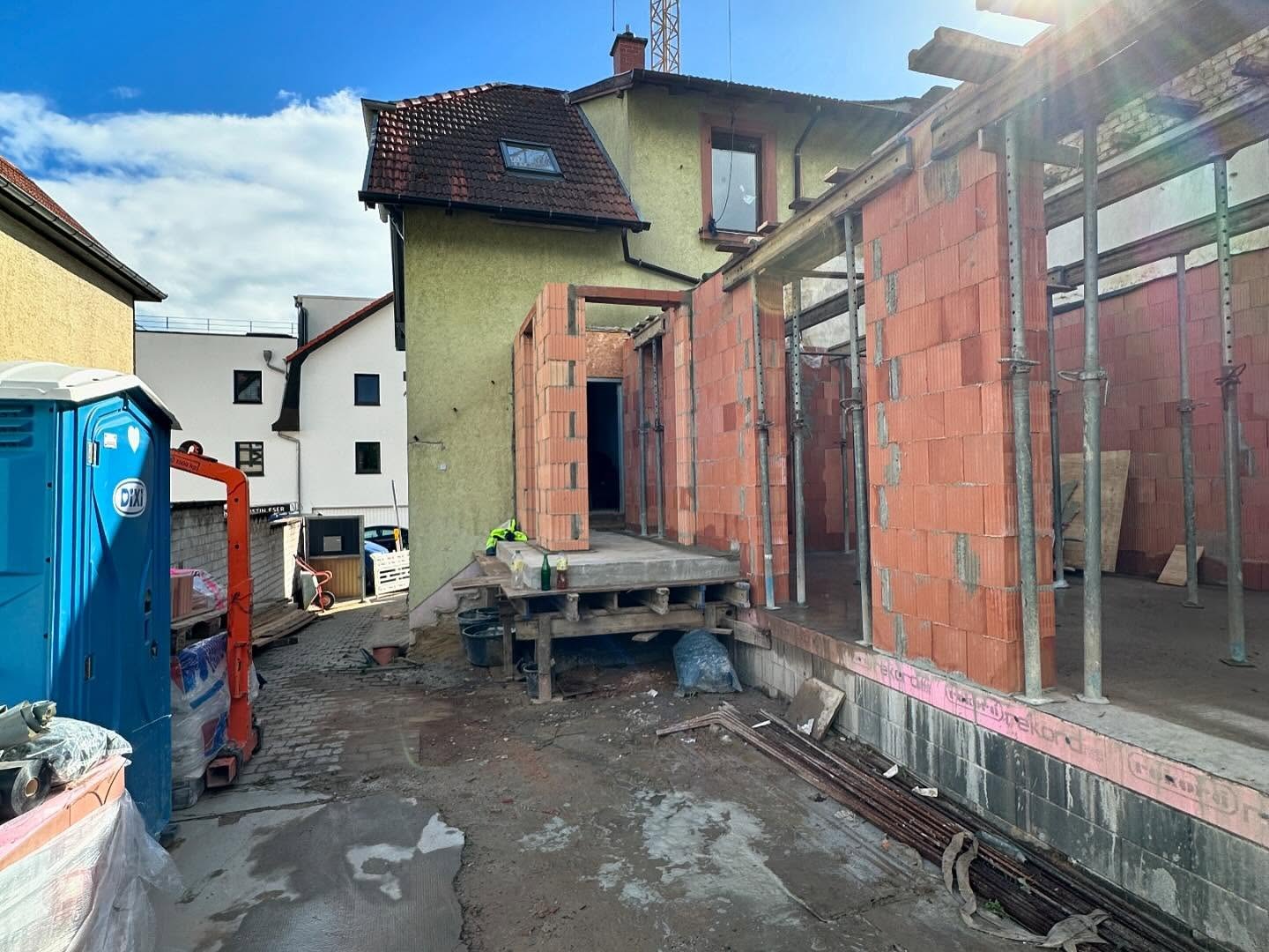 Rohbau des Anbaus mit hochgezogenen Poroton-Aussenwaenden, Tuersturz und auf Baustuetzen abgefangener Deckenschalung neben dem gruenen Altbau, links Dixi-Toilette und oranger Anhaengerkran bei blauem Himmel.