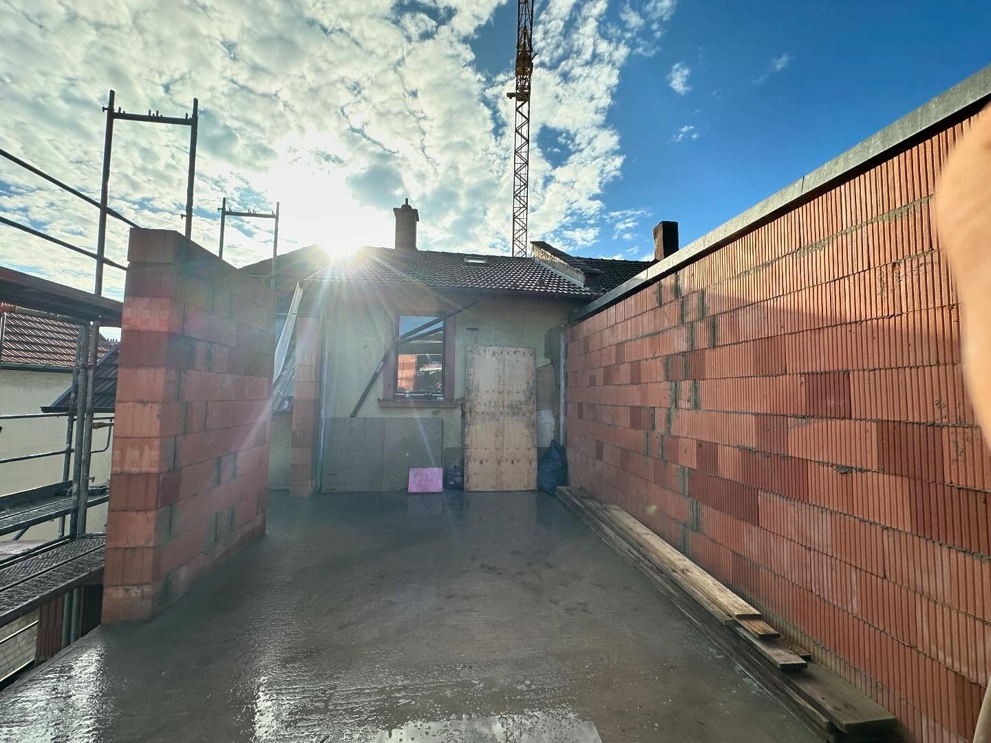 Gegenlichtaufnahme im Obergeschoss-Rohbau: Sonne bricht ueber das Dach des Bestandsgebaeudes, davor aufgemauerte Ziegelwaende, nasse Betondecke und ein Kranmast am Himmel
