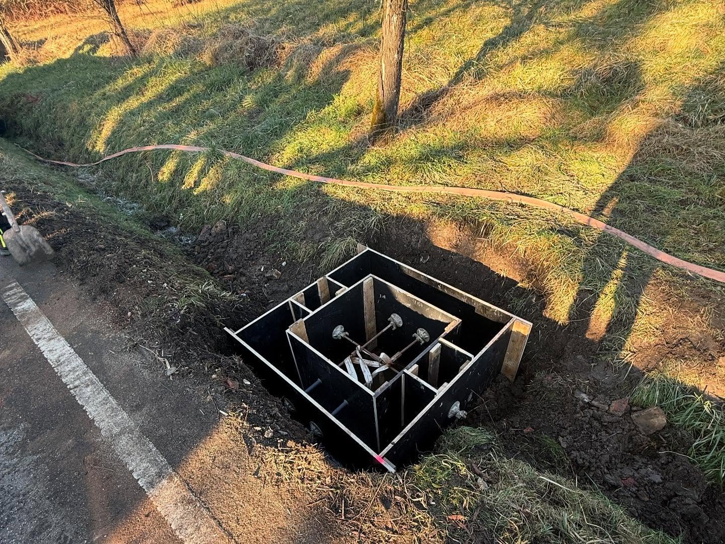 Draufsicht auf die fertig gestellte, exakt ausgesteifte schwarze Holzschalung eines quadratischen Ortbeton-Einlaufschachts in der Grabenböschung neben der Straße, im goldenen Abendlicht.