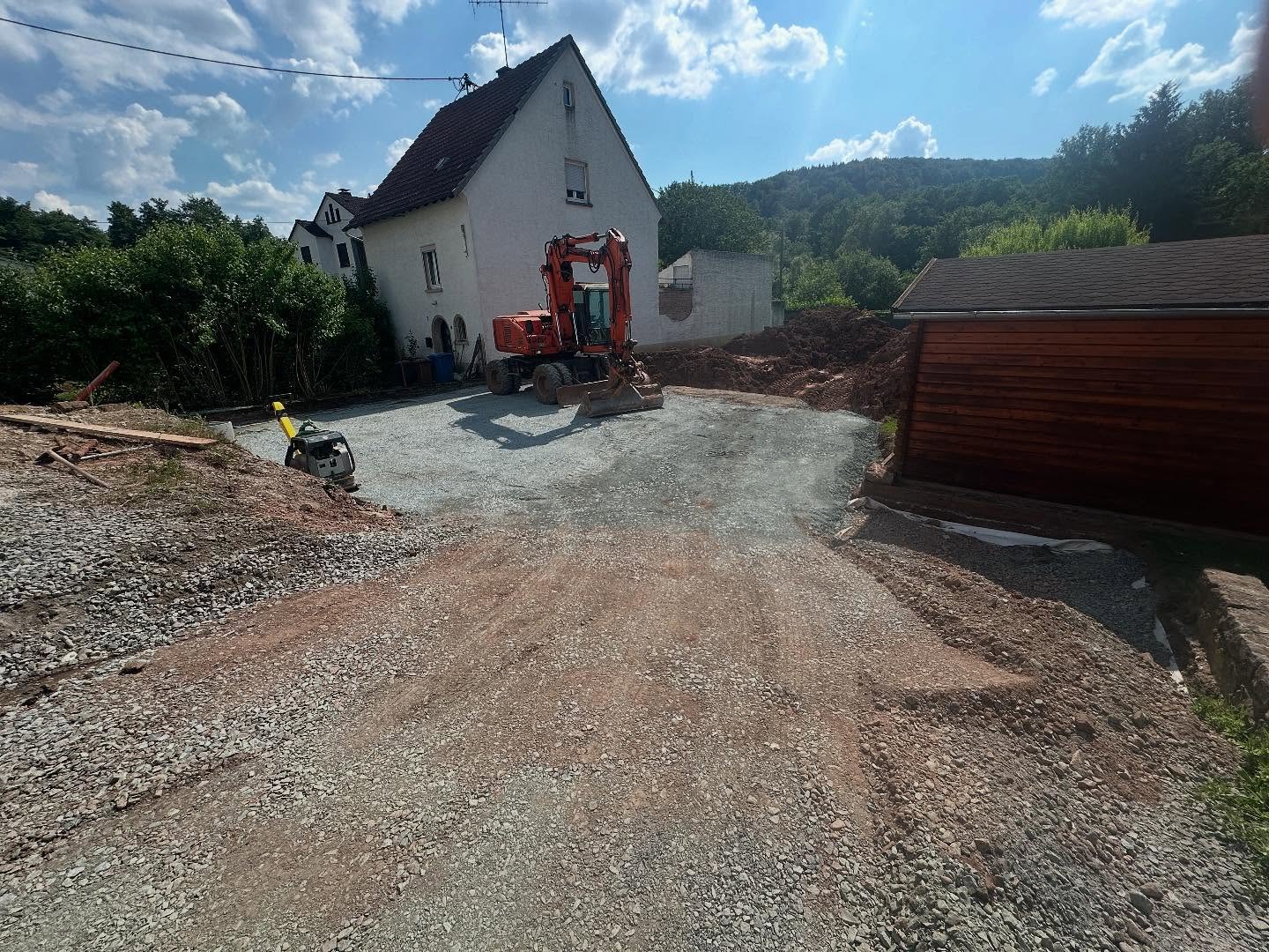 Fertig planierte und verdichtete Schotter-Tragschicht für Einfahrt und Garagenvorplatz mit orangem Mobilbagger und Rüttelplatte, im Hintergrund das weiße Wohnhaus und bewaldete Hügel unter blauem Himmel.