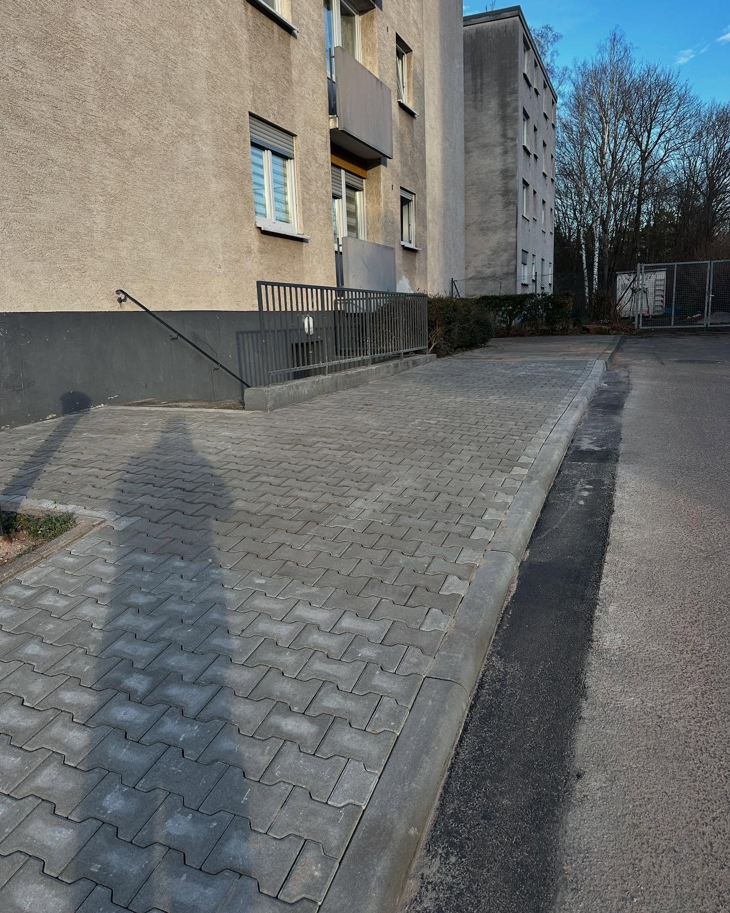 Neu verlegter Zufahrtsstreifen aus grauem Doppel-T-Verbundpflaster entlang der Sockelwand eines Mehrfamilienhauses, sauber mit durchlaufender Bordsteinkante zur Asphaltstrasse abgesetzt.