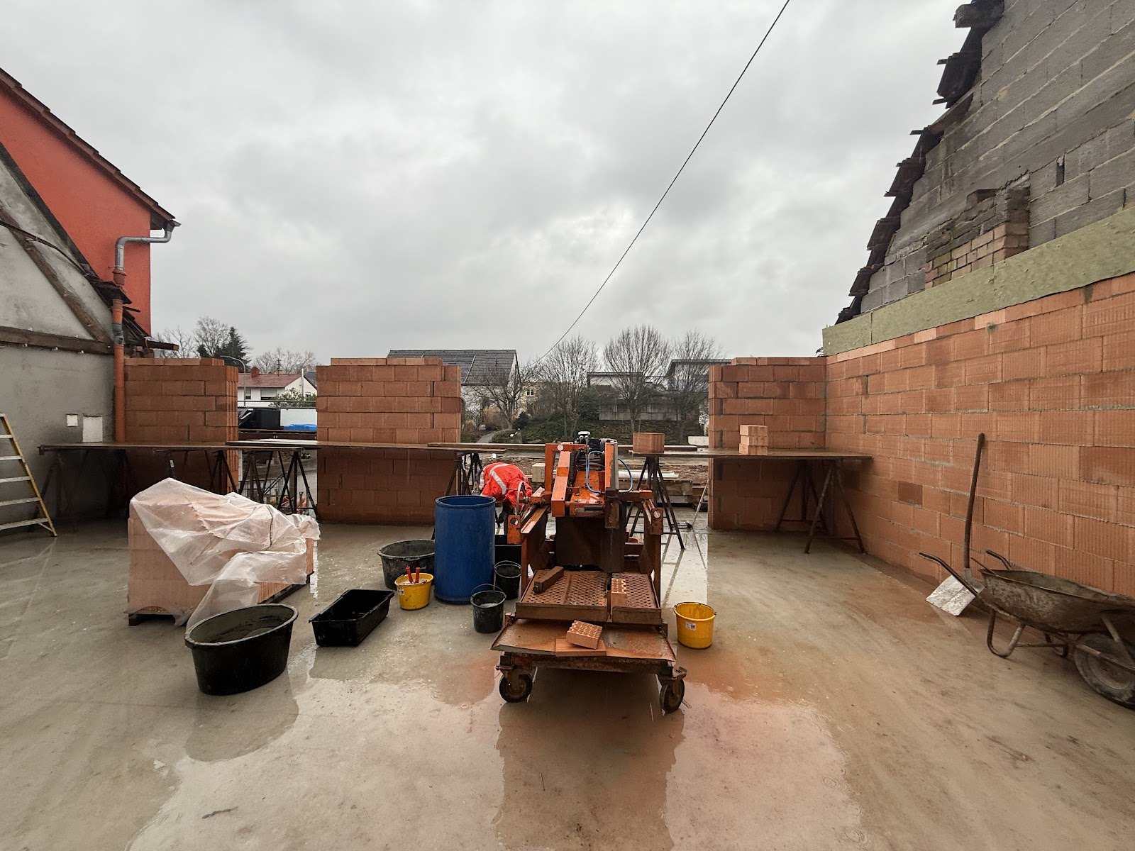 Aufgemauerte Ziegelwaende im Erdgeschoss einer Baulueckenbebauung mit Moertelkuebeln, Schubkarre und orangefarbenem Steinsaege-Wagen auf der frischen Betonsohle